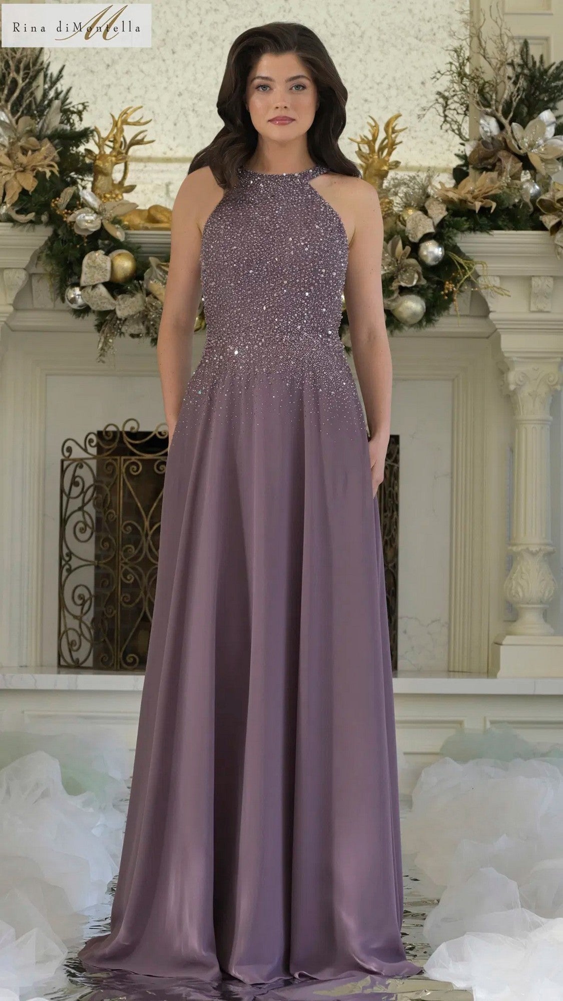 A woman stands before a decorated fireplace wearing the Rina Di Montella RD3304, a long formal dress featuring a sleeveless halter neckline, beaded bodice, and flowing purple chiffon A-line skirt.